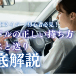車内でインストラクターが指を指しながら、20代女性ドライバーにハンドルの正しい持ち方を指導しているシーン。クロスハンドルと送りハンドルの操作を学べる記事のアイキャッチ画像。